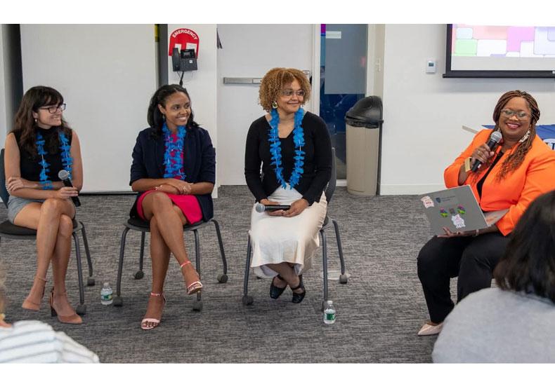 Four seated women in a classroom, one with a microphone speaking