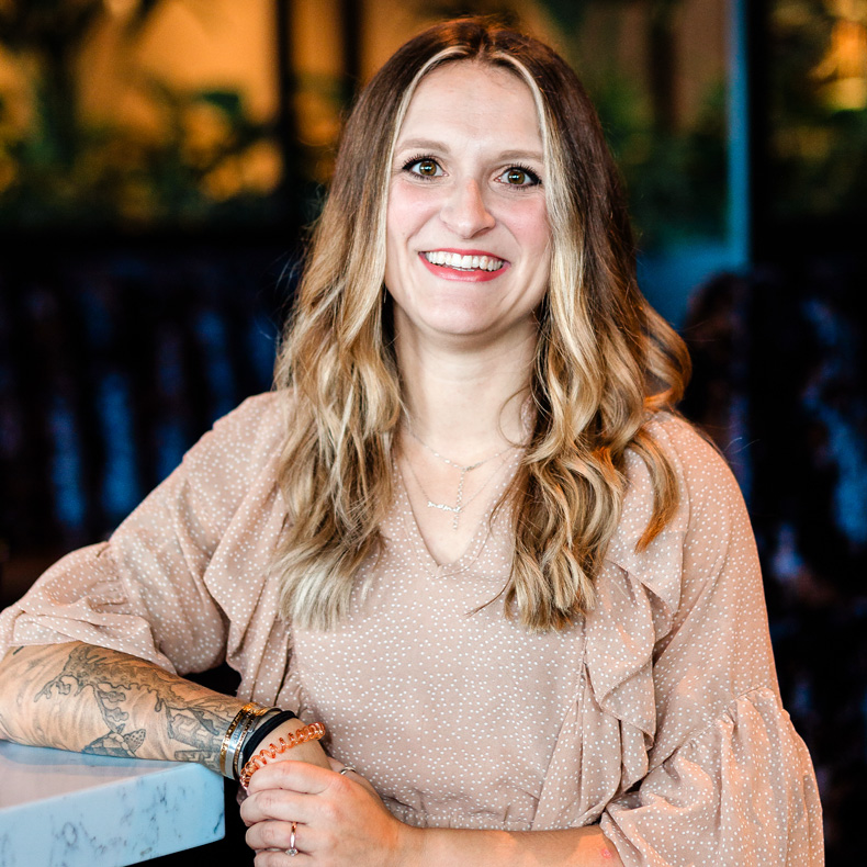 Smiling caucasian woman with blond hair, tan top, and tattoo on arm with dark background