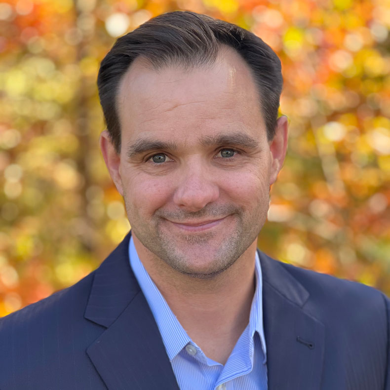 profile photo of smiling white male with short brown hair, button down shirt and navy jacket, with fall colors in the background