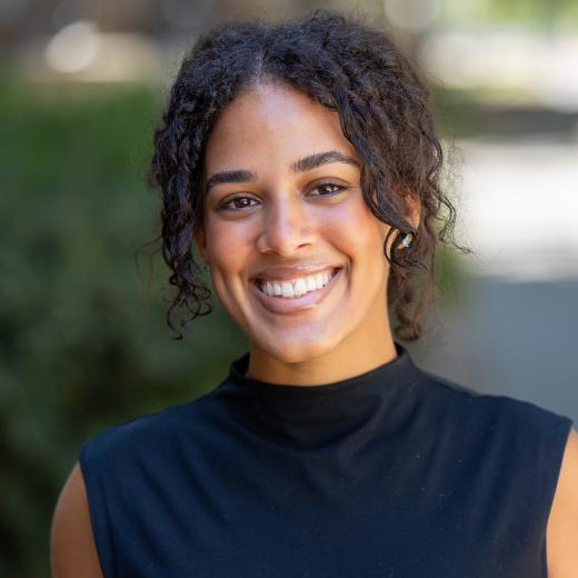 Portrait photo of female smiling with curly brown hair, dark blue sleeveless top, with blurred background