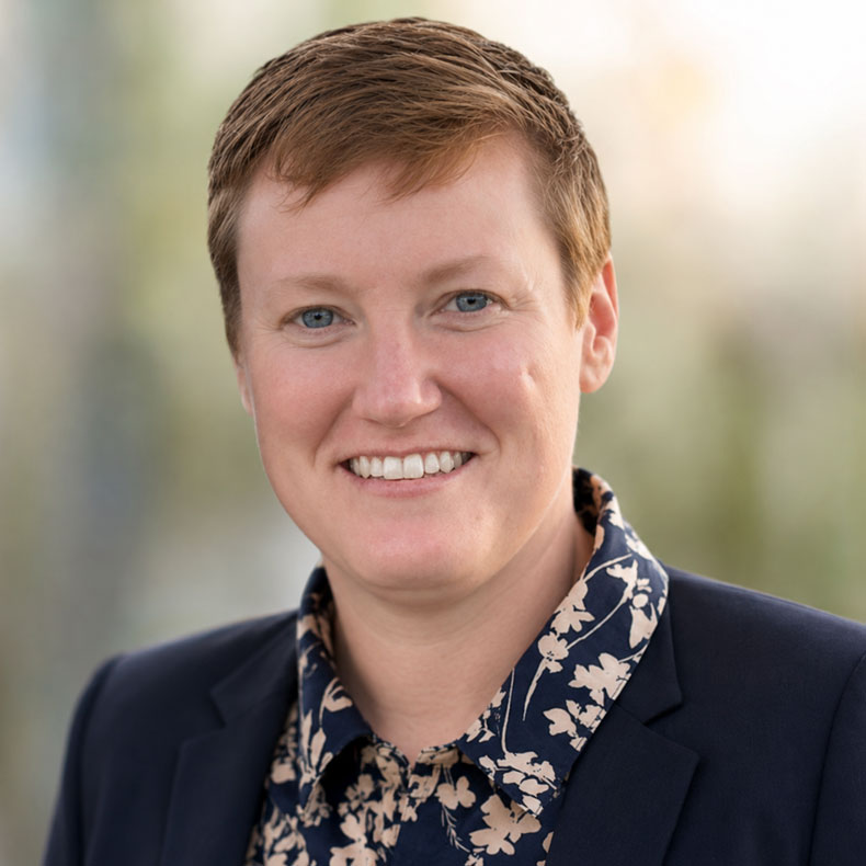 Portrait photo of smiling woman with short light red-brown hair, navy jacket, flowered shirt, and blurred background