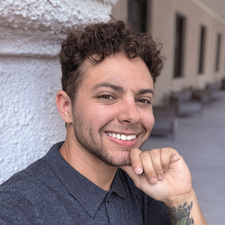 Portrait photo of smiling male, with hand to chin, curly brown hair, gray shirt, with a concrete column behind him and a blurred background