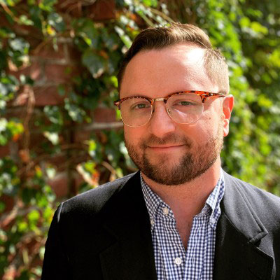 profile photo of smiling white male with short brown hair, glasses, button down shirt and jacket, with greenery in the background