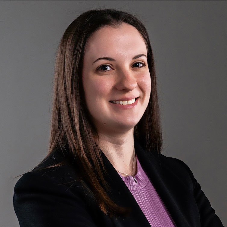 Portrait photo of smiling woman with long brown hair, lavender blouse, dark jacket on dark grey background