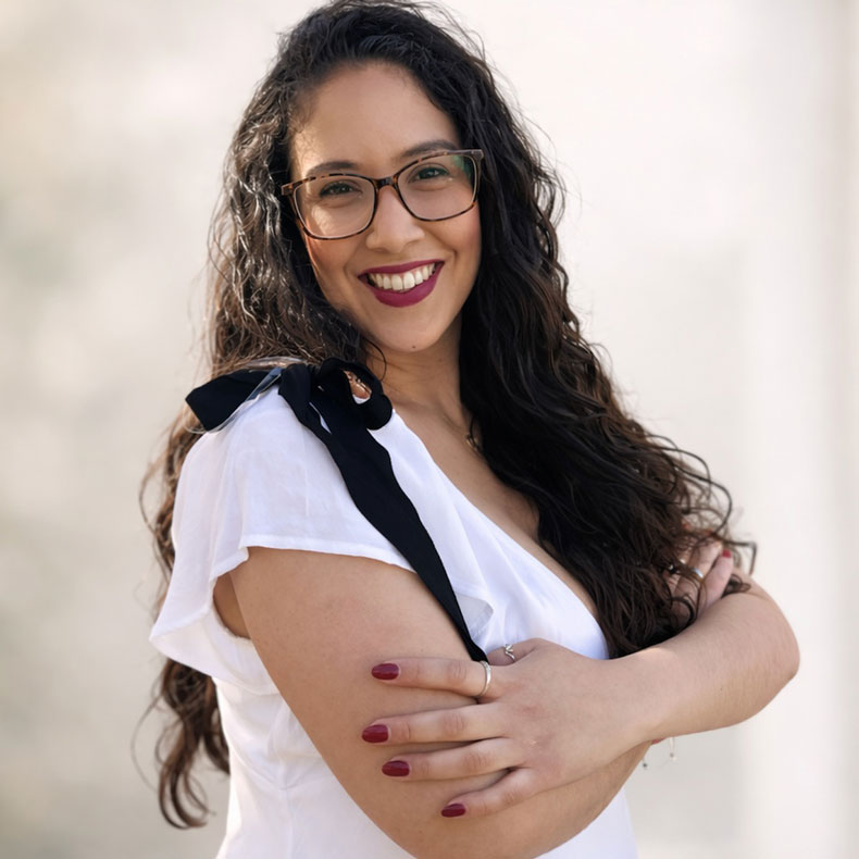 Portrait photo of smiling woman with long dark brown curly hair, glasses, white top, arms crossed in front of her, with a neutral backgeround