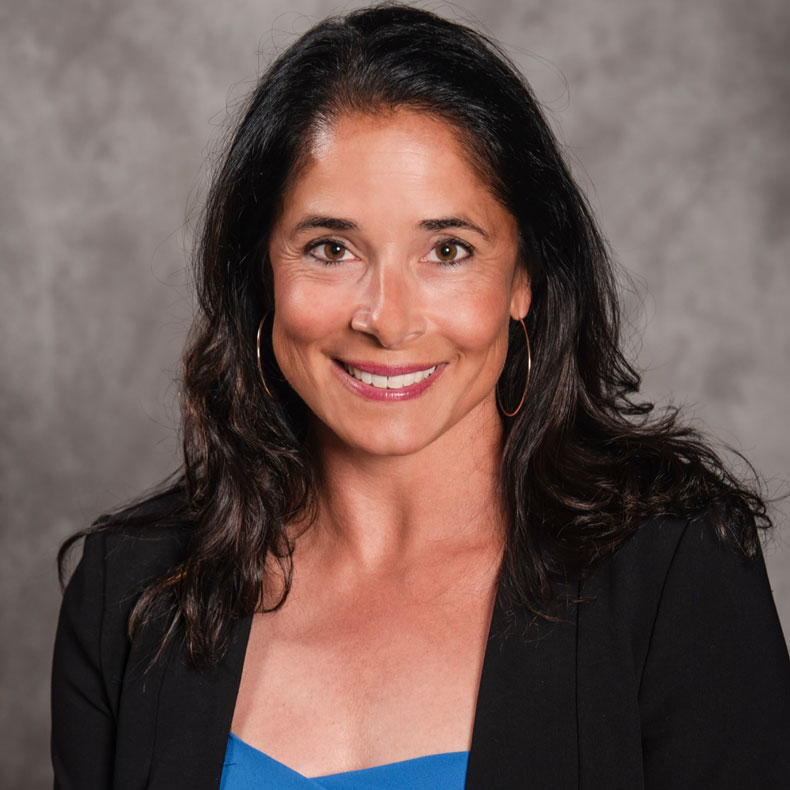 Profile photo of smiling woman with long brown hair, black jacket, blue blouse and neutral background
