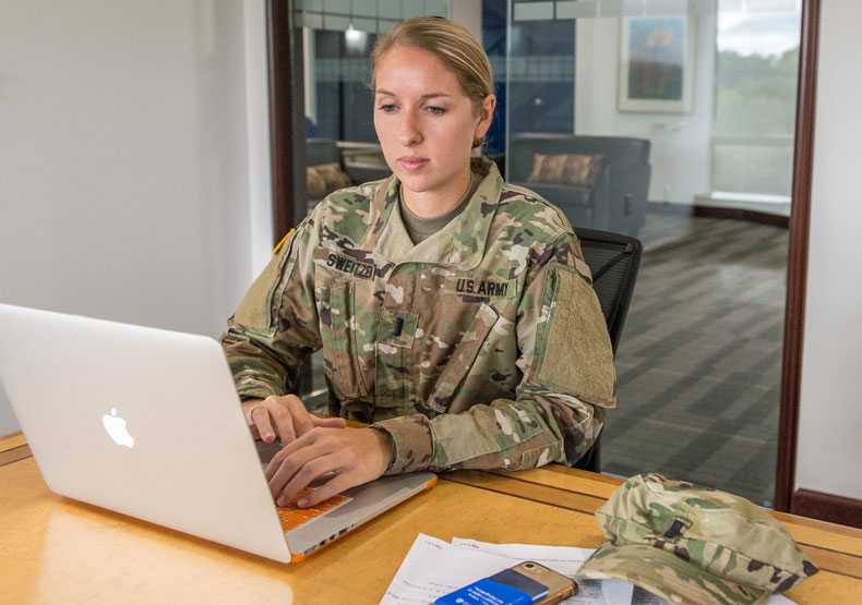woman in fatigues sitting at table typing into laptop