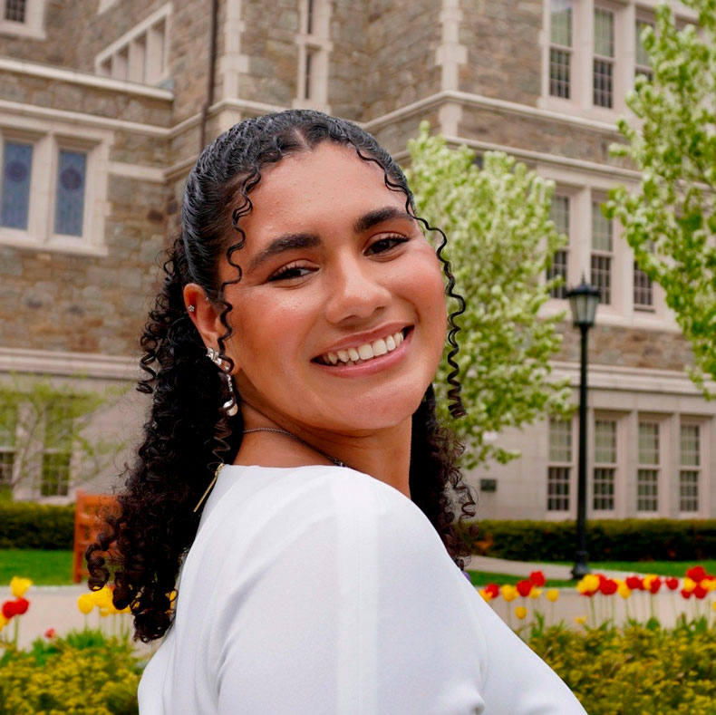 Close-up of woman smiling with brick building and flowers in the background