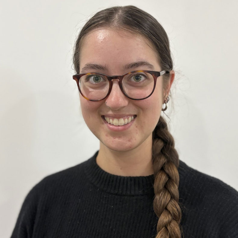 Portrait photo of smiling woman in glasses, with brown hair in a long braid, dark sweater and beige background