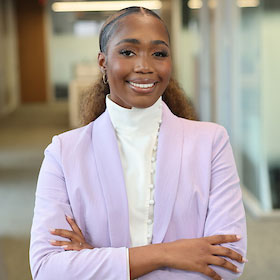 Photo of smiling woman, in a lavender jacket with blurred office environment behind her