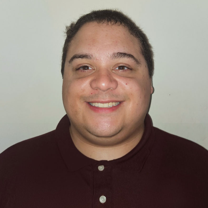 Portrait photo of smiling man with short dark hair, wearing a burgundy colored shirt, and a neutral background