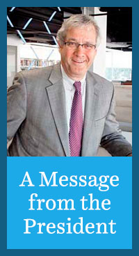 Image of man wearing gray coat and striped tie standing inside college building with text on bottom half