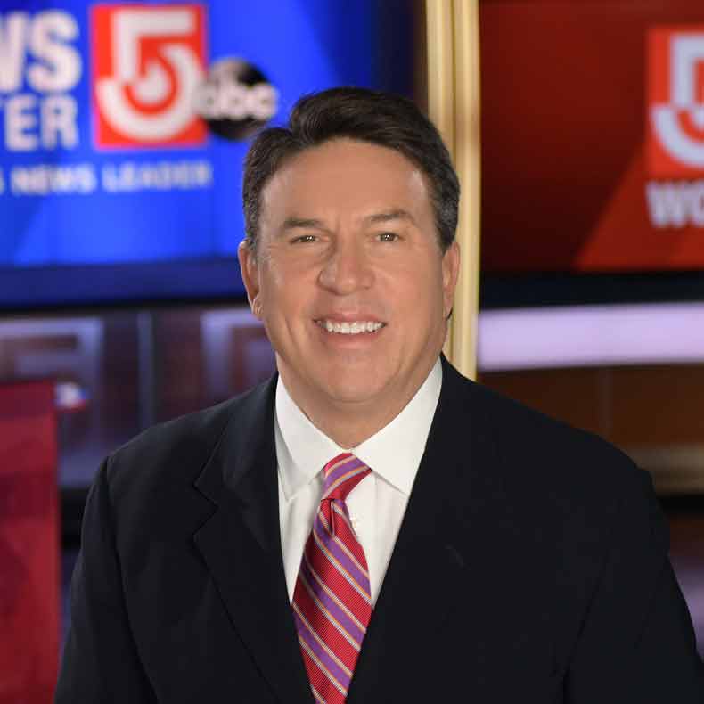 Photo of man in dark suit with TV logos behind him