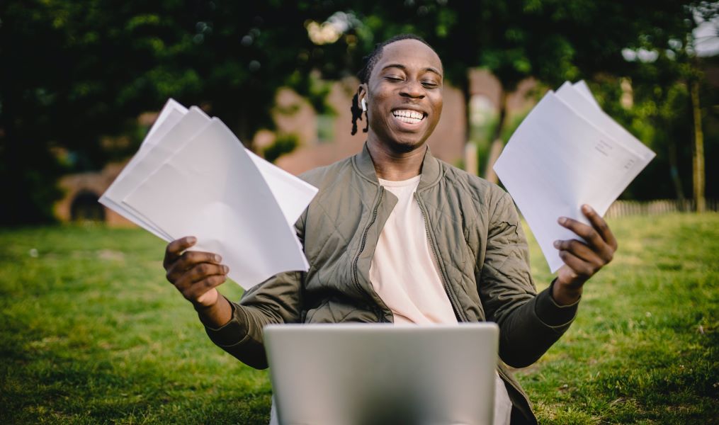 Smiling man sitting outside with a laptop, holding papers