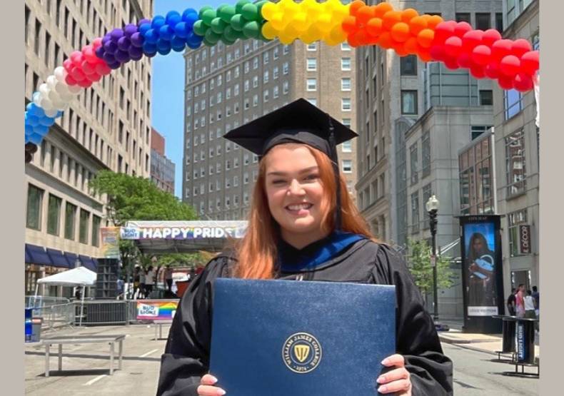 Colleen Deely, a young woman in a graduate's gown holding a diploma up beneath an arch of balloons in the streets of Boston