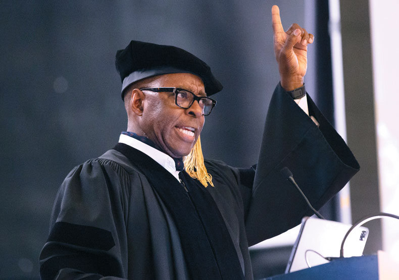 man in cap and gown speaking at podium with finger pointing towards sky