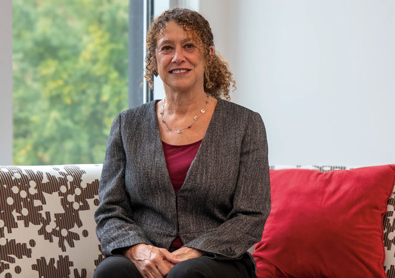 Woman in gray jacket sitting on a patterned couch with a window of greenery in the background