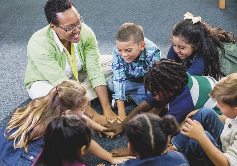 photo of group of smiling children with adult sitting in a circle with hands together in the middle