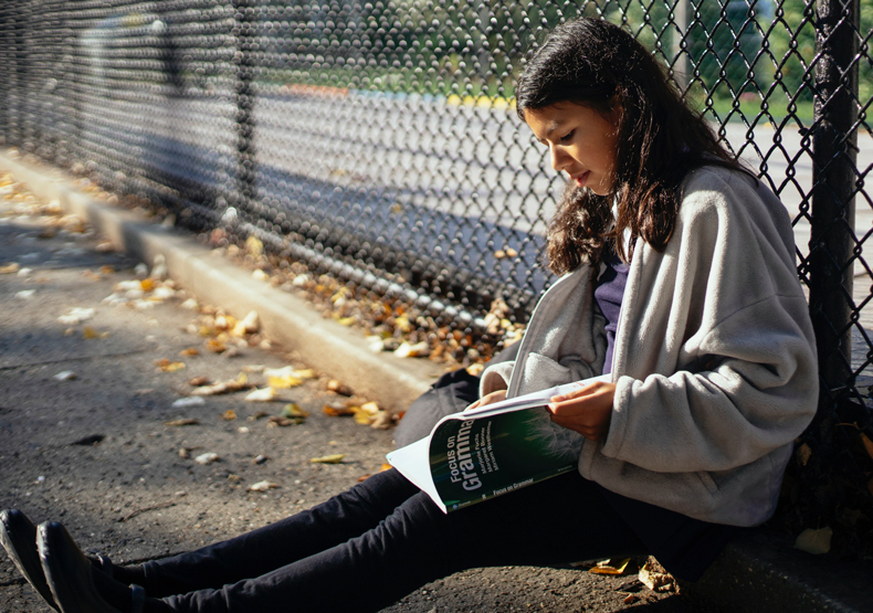 adolescent girl sitting next to chain link fence reading a book