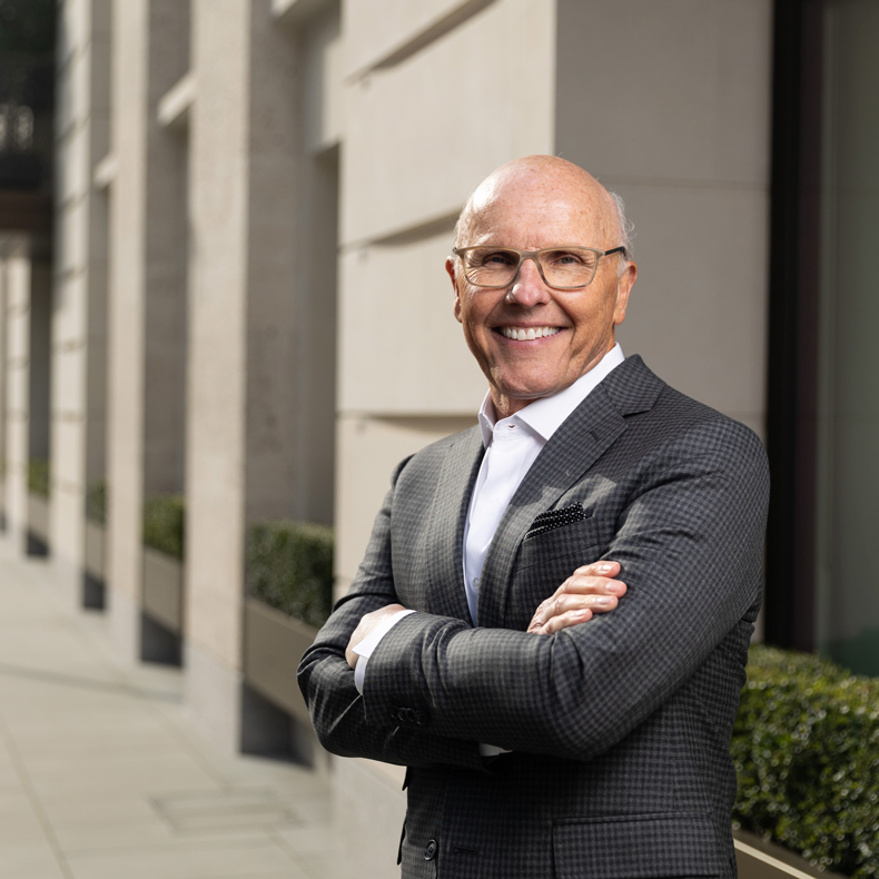 Photo of man smiling in in front of city building in gray suite with crossed arms and wearing glasses